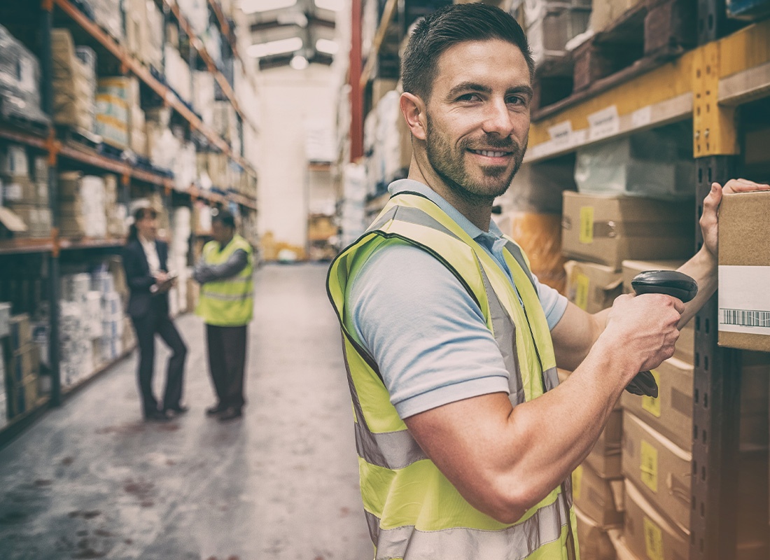 Warehousing and Logistics Insurance - Portrait of a Warehouse Worker Scanning a Box While Smiling With Fellow Colleagues Blurred in the Distance